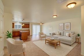 Williston apartment for rent - A bright open-concept living room here at Phoenix Ridge featuring a neutral sofa with accent pillows, a wooden coffee table, cozy upholstered chairs, and simple wall art, flowing into a kitchen with warm wood cabinetry, an island, tiled flooring, and sliding glass doors that bring in natural light.