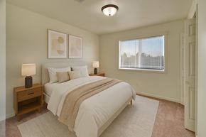 Williston apartment for rent - A bright neutral bedroom here at Phoenix Ridge featuring a comfortable bed with white bedding and a textured throw, matching wood nightstands with modern lamps, soft carpet flooring, minimalist wall art, and a large window with blinds that fills the space with natural light.