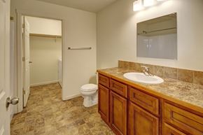 Williston apartment for rent - A clean neutral bathroom here at Phoenix Ridge featuring a wood vanity with a stone-look countertop and white sink, tile flooring in warm earth tones, a large wall mirror with overhead lighting, a toilet, and an adjacent walk-in closet with shelving and in-unit laundry access visible through the open doorway.