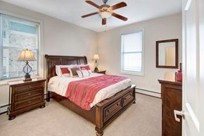 Williston apartment for rent - A cozy staged bedroom here at Prairie Pines at the Ridge featuring a dark wood bed with built-in storage drawers, neutral carpeting, matching nightstands with lamps, large windows that bring in natural light, and a ceiling fan that adds comfort and airflow.