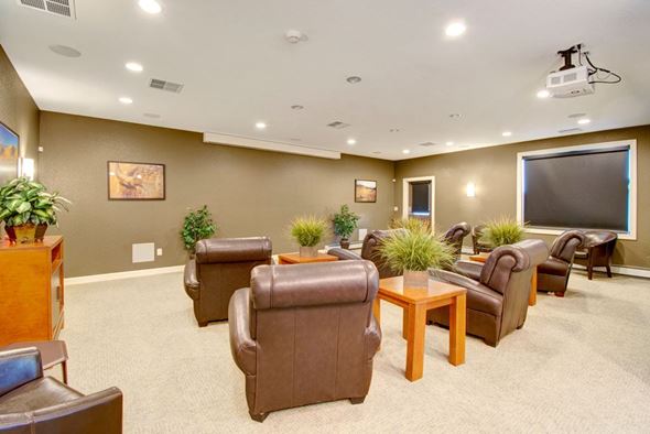 Williston apartment for rent - A comfortable resident lounge here at Prairie Pines at the Ridge featuring plush leather seating, wooden side tables with greenery, soft carpeted flooring, neutral accent walls, framed artwork, recessed lighting, and a ceiling-mounted projector creating a relaxed space for gatherings and entertainment.
