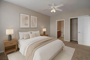 Williston apartment for rent - A serene bedroom here at Prairie Vista featuring neutral-toned walls, soft carpet flooring, a queen-sized bed with white bedding and a textured throw, matching bedside tables with lamps, modern abstract wall art, a ceiling fan with light, and direct access to a walk-in closet and ensuite bathroom.