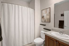 Williston apartment for rent - A clean bathroom here at Prairie Vista featuring a single-sink vanity with a light countertop, wood cabinetry, a large wall mirror, a toilet, floating wall shelves, neutral wall art, and a shower-tub combo with a white curtain and light tile flooring.