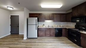 Williston apartment for rent - A spacious, modern kitchen here at Renaissance Heights featuring warm wood-style flooring, dark wood cabinetry, light countertops, a stainless steel refrigerator, black appliances, a double sink, and ample counter space with soft overhead lighting that creates a clean and functional cooking area.