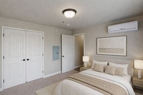 Williston apartment for rent - A bright, neutral bedroom here at Renaissance Heights featuring a comfortable bed with white bedding and beige accent pillows, matching bedside tables with lamps, soft carpet flooring, white double closet doors, and a wall-mounted air conditioning unit that creates a calm and inviting sleeping space.