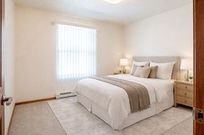 Williston apartment for rent - A bright, cozy bedroom here at Rockridge Apartments with beige carpet, soft neutral walls, and a large window fitted with vertical blinds that allow natural light to fill the room. A neatly made bed with light bedding is flanked by matching nightstands and lamps, creating a calm and inviting atmosphere.