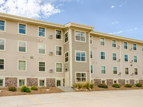Williston apartment for rent - A modern, multi-story apartment building exterior here at Rockridge Apartments with beige siding, white trim, and rows of large windows. The building features a central entry with steps, landscaped rock beds with greenery along the base, and a paved parking area in front under a clear blue sky.