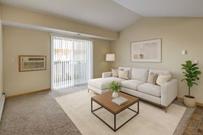 Williston apartment for rent - A bright, inviting living room here at Roosevelt East Apartments with beige carpet, soft neutral walls, and a sliding glass door with vertical blinds that brings in natural light. A light-colored sectional sofa sits on an area rug with a wooden coffee table, accented by simple wall art, a floor lamp, and a potted plant for a warm, comfortable feel.