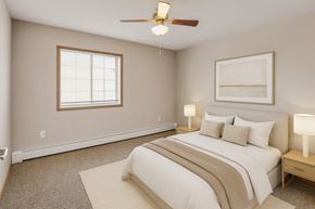 Williston apartment for rent - A calm, comfortable bedroom here at Roosevelt East Apartments with beige carpet, soft neutral walls, and a large window with blinds that lets in natural light. A ceiling fan with light hangs overhead, and the room is staged with a neatly made bed, matching nightstands, and lamps, creating a warm and relaxing atmosphere.