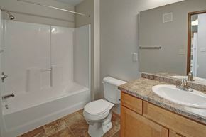 Williston apartment for rent - A clean, functional bathroom here at Roosevelt East Apartments with a bathtub and shower combo, neutral wall colors, and tiled flooring. A wooden vanity with a spacious countertop and sink sits beneath a large wall mirror, with a white toilet positioned nearby, creating a simple, well-lit, and easy-to-use layout.