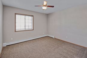 Williston apartment for rent - A spacious, empty bedroom here at Roosevelt East Apartments with beige carpet, soft neutral walls, and a window with blinds that brings in natural light. A ceiling fan with light is centered overhead, and the open layout with clean lines and wood-trim accents offers flexible space for a variety of bedroom furniture arrangements.