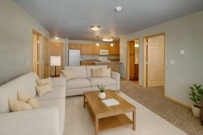 Williston apartment for rent - A warm, open living room here at Vue 28 Apartments with beige carpet, soft neutral walls, and coordinated wood trim throughout. A light-colored sectional sofa sits on an area rug with a wooden coffee table, while the open layout flows into the kitchen with wood cabinets, a breakfast bar, and overhead lighting, creating a comfortable, inviting space.