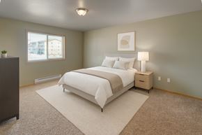 Williston apartment for rent - A bright, comfortable bedroom here at Vue 28 Apartments with beige carpet, soft neutral walls, and a large window with blinds that brings in natural light. The room features a neatly made bed with light bedding, a bedside table with a lamp, simple wall art, and an open layout that feels calm and inviting.