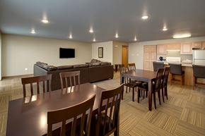 Williston apartment for rent - A spacious community gathering area here at Vue 28 Apartments featuring multiple wooden dining tables with chairs, a comfortable seating area with sofas, and an open kitchen with a breakfast bar and cabinetry. Recessed ceiling lights and patterned flooring create a bright, welcoming space designed for socializing, dining, or group activities.