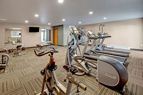 Williston apartment for rent - A well-equipped fitness center here at Vue 28 Apartments featuring cardio machines, weight equipment, and exercise benches arranged across a spacious, open floor plan. The room includes mirrored walls, wall-mounted televisions, patterned flooring, and bright overhead lighting, creating a comfortable and functional space for daily workouts.