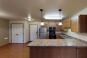 Williston apartment for rent - A spacious kitchen here at Williston Garden Apartments with warm wood cabinets, wood-style flooring, and expansive countertops that include a large island for added prep and seating space. The kitchen features a refrigerator, stove, microwave, and double sink, with pendant lighting and an open layout that connects easily to the surrounding living area.