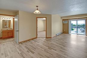 Williston apartment for rent - A spacious, open living area here at Williston Garden Apartments with wood-style flooring, soft neutral walls, and an airy layout. A sliding glass door with vertical blinds leads to a private balcony and allows natural light to fill the room, while baseboard heating, overhead lighting, and nearby access to the bedroom and bathroom add comfort and convenience.