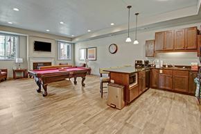 Williston apartment for rent - A stylish community lounge here at Williston Garden Apartments with wood-style flooring, recessed lighting, and a welcoming open layout. A pool table anchors the seating area near a wall-mounted television and fireplace, while a full kitchen with wood cabinets, an island, and pendant lighting creates a comfortable space for gatherings and social events.