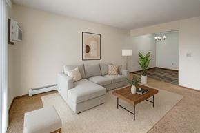 Williston apartment for rent - A cozy living room here at Windscape Apartments featuring plush carpet, soft neutral walls, and a modern sectional sofa arranged on a textured area rug. A wooden coffee table, simple wall art, and potted greenery add warmth, while an open view into the dining area creates a comfortable, inviting layout.