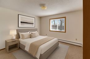 Williston apartment for rent - A calm, well-appointed bedroom here at Windscape Apartments featuring soft neutral walls, plush carpet, and a large window that provides natural light and outdoor views. A neatly styled bed with a padded headboard, coordinating nightstand, and simple artwork creates a comfortable, restful sleeping space.