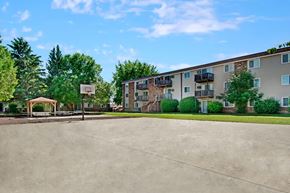 Williston apartment for rent - A bright outdoor recreation area here at Windscape Apartments featuring a large open concrete court with a basketball hoop, surrounded by green lawns, mature trees, and nearby apartment buildings with private balconies, creating a welcoming community space for residents to enjoy outdoor activities and social gatherings.