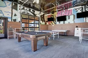 Tulsa apartment for rent - A lively game room here at 71 at Tulsa Hills featuring a pool table, ping pong table, cornhole boards, mounted TVs, and bold mural artwork, creating a fun and social space for residents to relax and gather.