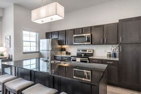 Owasso apartment for rent - A sleek kitchen here at 98 Apartments with dark cabinetry, stainless steel appliances, glossy granite countertops, a large island with seating, and bright lighting, creating a modern and polished space for cooking and entertaining.