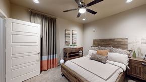 Owasso apartment for rent - A warm bedroom here at 98 Apartments with soft carpet, a wood-accent bed frame, neutral bedding with patterned pillows, a ceiling fan, and floor-length curtains, creating a cozy and restful retreat.