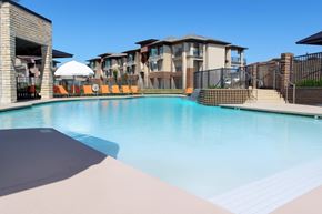 Owasso apartment for rent - Here at 98 Apartments, a resort-style outdoor pool with clear blue water, modern stone accents, bright orange lounge chairs, and surrounding apartment buildings that create a relaxing and upscale atmosphere.
