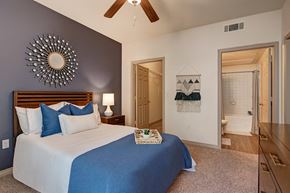 Oklahoma City apartment for rent - A cozy bedroom here at Aviare Arts District with soft carpet, a blue accent wall, a comfortable bed with neutral and blue bedding, a ceiling fan, and an attached bathroom visible, creating a calm and inviting retreat.
