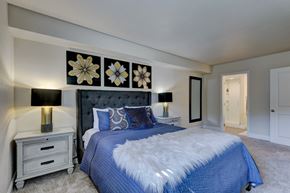 Oklahoma City apartment for rent - A comfortable bedroom here at Indigo with soft carpet flooring, neutral walls, and bedside lighting. A spacious bed with plush bedding and decorative wall art creates a relaxing and inviting atmosphere, while a nearby doorway leads to an attached bathroom.