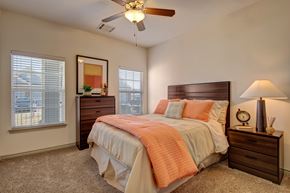 Oklahoma City apartment for rent - Here at Liberty Pointe, a bright bedroom with warm neutral walls and soft carpet, featuring a wooden bed frame styled with a white bedspread and standout coral bedding accents. Matching wood nightstand and dresser add warmth, while a table lamp, framed wall art, and natural light from two windows create a clean, comfortable feel.
