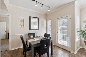 Oklahoma City apartment for rent - A dining area here at Lincoln at Central Park featuring a dark wood dining table paired with four black upholstered chairs, set on warm wood-style flooring. Natural light enters through a glass patio door and side window with white blinds, while a wall mirror, decorative wall panels, and a simple candle centerpiece create a clean, welcoming, and comfortable space.