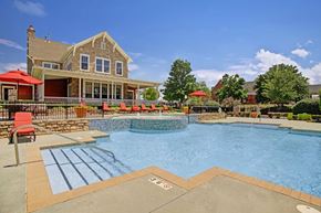 Tulsa apartment for rent - A resort-style pool here at Memorial Creek featuring clear blue water, stone accents, red lounge chairs with umbrellas, wide deck seating, and a bright clubhouse backdrop under a sunny sky.