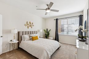 Oklahoma City apartment for rent - A bedroom here at Quail Landing with soft carpet, a light upholstered bed, white bedding with a mustard accent pillow, matching side tables with lamps, a ceiling fan, and natural light through wide windows with dark curtains.