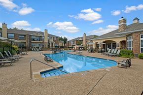 Oklahoma City apartment for rent - A resort-style pool area here at Quail Landing with clear blue water, tan pool decking, black lounge chairs, shaded seating, and surrounding brick apartment buildings under a bright blue sky.