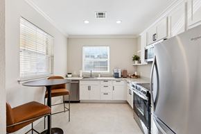 Oklahoma City apartment for rent - A kitchen here at Quail Landing with white cabinets, stainless steel appliances, light tile flooring, a small round dining table with brown leather stools, bright windows with blinds, and a clean, modern layout.