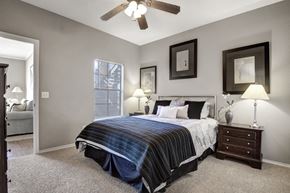 Oklahoma City apartment for rent - A comfortable bedroom here at Stoneleigh on May with beige carpet, soft gray walls, and a ceiling fan with light. A large bed with neutral bedding is flanked by matching nightstands, while a window with blinds brings in natural light.