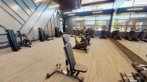 Yukon apartment for rent - A fitness center here at Trailwinds with modern cardio and strength machines, patterned flooring with subtle orange accents, mirrored walls, high ceilings, and large windows bringing in natural light.