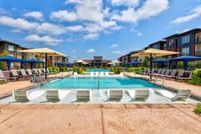 Yukon apartment for rent - A resort-style pool here at Trailwinds with a shallow lounging ledge, white in-water chaise loungers, blue umbrellas, surrounding lounge chairs, and landscaped courtyards framed by modern apartment buildings under a bright blue sky.