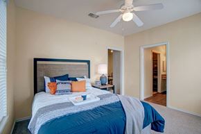 Broken Arrow apartment for rent - A bedroom here at Woodland Park with a beige color palette, white ceiling fan, and carpeted floor, featuring a wood-tone headboard bed with blue and orange accent pillows, a bedside lamp, and open doorways to a closet and bath area.