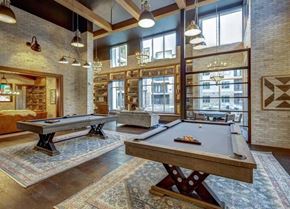 Cedar Park apartment for rent - A game room here at Alden at Cedar Park with two pool tables, patterned rugs, tall windows, brick walls, wood beams, and warm pendant and chandelier lighting in a spacious lounge.