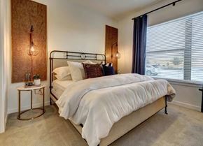 Cedar Park apartment for rent - A bedroom here at Alden at Cedar Park with a black metal bed frame, white bedding, dark accent pillows, blue curtains, round gold side table, and warm wall panels with hanging lights.