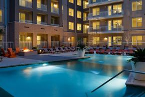 Dallas apartment for rent - A resort-style pool courtyard here at Arpeggio with a lit swimming pool, lounge chairs, a fire pit seating area, and surrounding apartment balconies glowing with warm evening lighting.