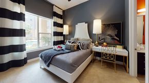 Dallas apartment for rent - A stylish bedroom here at Arpeggio with a white upholstered bed, gray bedding, bold black and white striped curtains, dark accent wall, gold and white desk, and a large window with bright natural light.