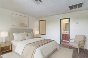 Midland apartment for rent - A comfortable bedroom here at Avanti Townhomes with plush carpet and soft neutral walls, featuring a cozy bed with light bedding, matching nightstands and lamps, a seating chair, and an attached bathroom visible through an open doorway for a calm and inviting retreat.