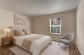 Midland apartment for rent - A bright bedroom here at The Bradford with beige carpet, soft neutral walls, and a large window with blinds allowing natural light to fill the space. A neatly staged bed with neutral bedding and a simple accent chair create a calm, comfortable, and inviting atmosphere.