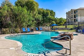 Austin apartment for rent - A resort-style pool here at Canyon Vistas with a curved swimming pool, stone water feature, blue lounge chairs, shaded seating, and surrounding greenery in a relaxing outdoor setting.