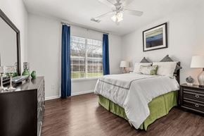 Austin apartment for rent - A bright bedroom here at Canyon Vistas with wood-style flooring, a white bed with green accent bedding, bedside tables with lamps, a large window with blue curtains, and a ceiling fan.