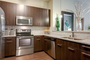 Dallas apartment for rent - A kitchen here at Cityplace Heights with dark wood cabinets, stainless steel appliances, white stone counters, tile backsplash, double sink, and green glass decor accents by the pass-through opening., encompassing a warm and open space.